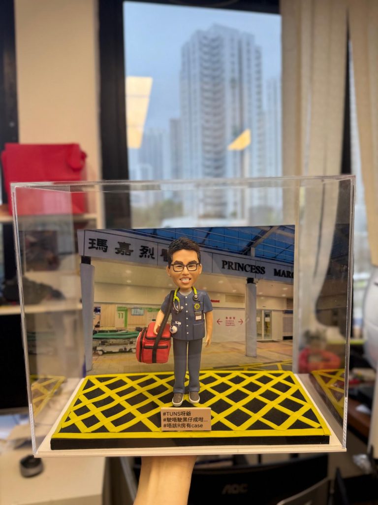 訂製醫生人像公仔｜醫護人員穿著深藍色醫護制服，胸前掛著聽診器，手持紅色醫療急救袋，背景為香港「瑪嘉烈醫院 Princess Margaret Hospital」正門場景。此公仔以急症室醫生為原型，作為專屬醫護禮物與紀念擺設。