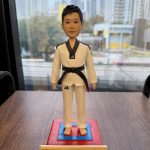 一個穿著白色跆拳道服、佩戴黑帶的人像公仔，站在紅藍色跆拳道比賽場地上。公仔有著短髮，面容堅毅，表現出一種積極的精神。