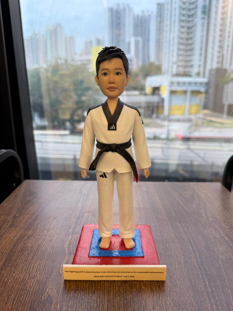 一個穿著白色跆拳道服、佩戴黑帶的人像公仔，站在紅藍色跆拳道比賽場地上。公仔有著短髮，面容堅毅，表現出一種積極的精神。