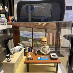 訂製醫生人像公仔｜教授坐於辦公桌前，桌面擺放電腦滑鼠、筆、紙張與個人照片，背景還原窗外大學醫院景觀，桌邊配有醫療監測儀器與實驗設備，展現醫護專業氛圍與退休紀念禮物的獨特價值。