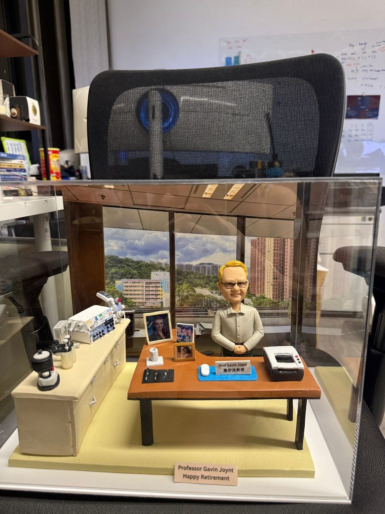 訂製醫生人像公仔｜教授坐於辦公桌前，桌面擺放電腦滑鼠、筆、紙張與個人照片，背景還原窗外大學醫院景觀，桌邊配有醫療監測儀器與實驗設備，展現醫護專業氛圍與退休紀念禮物的獨特價值。