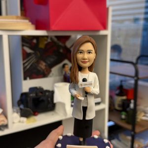手掌托起的記者主題人像公仔：長髮女記者穿白色外套與黑色長褲，右手持麥克風、左手拿稿件，底座為深紫色圓形舞台並點綴白色燈泡，前方附有日期與字樣的小銘牌；背景為辦公室書架與攝影器材。整體呈現專業而溫暖的致敬氛圍，適合作為記者人像公仔榮休禮物與收藏擺設。