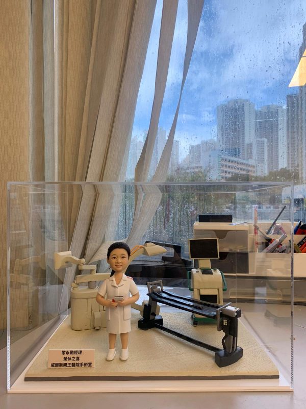 護士人像公仔，身穿白色制服，手持文件，站在醫療手術室場景中。背景包含手術燈、醫療監測儀與手術台等專業醫療設備，整體設計逼真，象徵醫護人員的專業與關懷。此公仔是專為醫生或護士設計的客製化禮物，適合作為醫護感謝、升職或退休紀念。