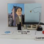 一款以「授人以漁」為主題的人像公仔：短髮男子穿西裝外套、淺綠色領帶與黑長褲，站在白色地台上，前方放有名牌，上寫「Samuel Chan 恩賜 授人以漁」。背景板左側為建築物照片，右側為淡藍色面板，上有黑色釣線示意；場景中擺放一張黑色治療/按摩床與工具箱，右前方立有灰色釣竿與紅色小魚模型；地台前緣有「Cutefigure_hk」字樣。整體呈教育與分享精神的釣魚人像公仔與人像公仔。