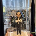 一尊以警務處「警長」職級為主題的人像公仔置於透明壓克力展示盒中，由左手掌托起近距離拍攝。公仔五官細緻：短而整齊的黑髮、劉海向上梳理；配戴圓框眼鏡，露出齒列微笑。上身著白色立領襯衫，袖口整潔收邊；外搭黑色無袖戰術背心，面料呈細霧面質感。左胸配有長條形識別牌與織章貼片，右胸口袋蓋上有淺色方形證章飾片與小型徽記；背心立體口袋、門襟與下擺車縫線條清晰。下身為深灰長褲，褲線筆直，腳穿黑色皮鞋，鞋面有亮面反光。公仔雙臂在胸前自然交疊，手掌比例圓潤，呈沉著自信的站姿。腳下為米白色粗紋理台座，前緣安置金色金屬名牌，刻有「祝賀…警長 榮陞之喜」等字樣（其餘字樣略）。展示盒後方插入高樓林立的商業街景背景板，仰角視角可見玻璃帷幕反射雲影與樹梢。更外層透過辦公室落地窗可見實景住宅高樓群與藍天，窗面映出長條燈具反光；畫面左側下方可見毛絨外套袖口與桌面書籍、文具與收納盒等物件，強化在職場環境中呈交禮的情境。