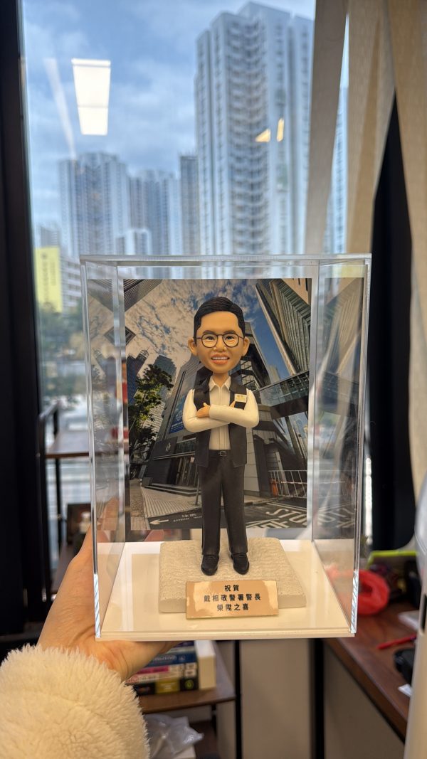 一尊以警務處「警長」職級為主題的人像公仔置於透明壓克力展示盒中，由左手掌托起近距離拍攝。公仔五官細緻：短而整齊的黑髮、劉海向上梳理；配戴圓框眼鏡，露出齒列微笑。上身著白色立領襯衫，袖口整潔收邊；外搭黑色無袖戰術背心，面料呈細霧面質感。左胸配有長條形識別牌與織章貼片，右胸口袋蓋上有淺色方形證章飾片與小型徽記；背心立體口袋、門襟與下擺車縫線條清晰。下身為深灰長褲，褲線筆直，腳穿黑色皮鞋，鞋面有亮面反光。公仔雙臂在胸前自然交疊，手掌比例圓潤，呈沉著自信的站姿。腳下為米白色粗紋理台座，前緣安置金色金屬名牌，刻有「祝賀…警長 榮陞之喜」等字樣（其餘字樣略）。展示盒後方插入高樓林立的商業街景背景板，仰角視角可見玻璃帷幕反射雲影與樹梢。更外層透過辦公室落地窗可見實景住宅高樓群與藍天，窗面映出長條燈具反光；畫面左側下方可見毛絨外套袖口與桌面書籍、文具與收納盒等物件，強化在職場環境中呈交禮的情境。