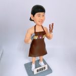 燒烤主題人像公仔：戴黑色頭巾的師傅身穿棕色圍裙與白色運動鞋，右手握兩串烤肉串，左手持金屬夾；站在灰色石磚紋底座上，前方白色小立牌寫有「串燒聖手」字樣。公仔面帶微笑、肌肉線條分明，整體呈現路邊攤／燒烤店職人的活力氛圍，適合作為燒烤師傅生日禮物與店內展示。