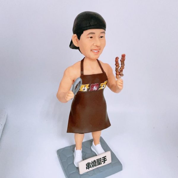 燒烤主題人像公仔：戴黑色頭巾的師傅身穿棕色圍裙與白色運動鞋，右手握兩串烤肉串，左手持金屬夾；站在灰色石磚紋底座上，前方白色小立牌寫有「串燒聖手」字樣。公仔面帶微笑、肌肉線條分明，整體呈現路邊攤／燒烤店職人的活力氛圍，適合作為燒烤師傅生日禮物與店內展示。