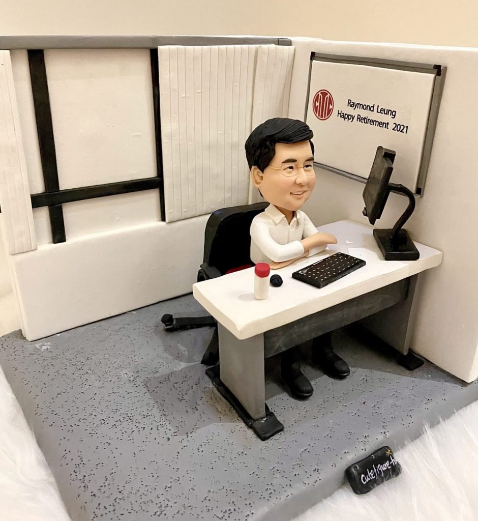 一座辦公室情境的人像公仔：短髮戴眼鏡的男子穿白襯衫與黑皮鞋，坐在黑色辦公椅上，雙臂自然靠在白色長方形辦公桌。桌上擺有單螢幕顯示器（黑色可調臂座）、長條鍵盤、滑鼠與一枚紅白公章。右後方白色看板印有圓形紅色標記與「Raymond Leung Happy Retirement 2021」致意字樣；周圍以白色隔板與灰色地台營造開放式隔間辦公室的氛圍。整體呈現簡潔沉穩的職場與榮退紀念場景。