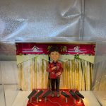 透明展示盒內的一款定製人像公仔：背景為紅底金字「××酒家」拱門與垂幕，象徵中式酒樓／餐館；公仔塑成中年女店主形象，短髮、笑容親切，穿紅色繡花上衣配黑長褲與涼鞋，右手持麥克風作開幕致辭。腳下紅色小舞台點綴暖黃燈串，前方陳列多個黑色音響／麥克風座，整體呈現宴會式開張氛圍。台前示例字牌寫「生日快樂」，實際下單可改為店名與開業賀詞，適合作為店鋪開張、新店啟業的人像公仔禮物。