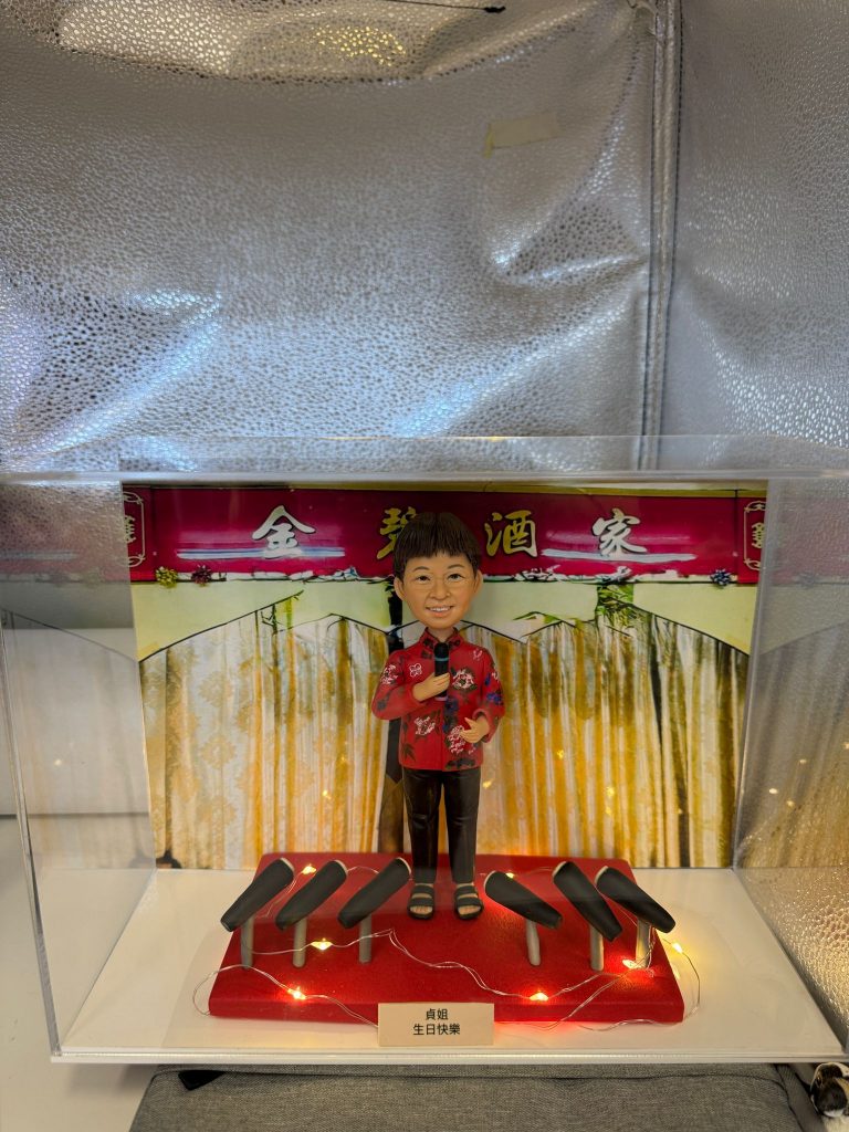 透明展示盒內的一款定製人像公仔：背景為紅底金字「××酒家」拱門與垂幕，象徵中式酒樓／餐館；公仔塑成中年女店主形象，短髮、笑容親切，穿紅色繡花上衣配黑長褲與涼鞋，右手持麥克風作開幕致辭。腳下紅色小舞台點綴暖黃燈串，前方陳列多個黑色音響／麥克風座，整體呈現宴會式開張氛圍。台前示例字牌寫「生日快樂」，實際下單可改為店名與開業賀詞，適合作為店鋪開張、新店啟業的人像公仔禮物。