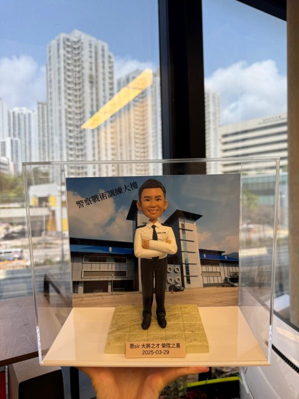 一款為香港警務人員訂製的晉升人像公仔，公仔身穿白色警官制服，背景為警察戰術訓練大樓，用以祝賀其榮陞之喜。
