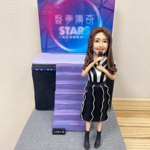 一位女歌手的人像公仔，她身穿黑白間條表演服，手持咪高峰，站在一個印有「聲夢傳奇 STARS ACADEMY」標誌的紫色舞台佈景前。