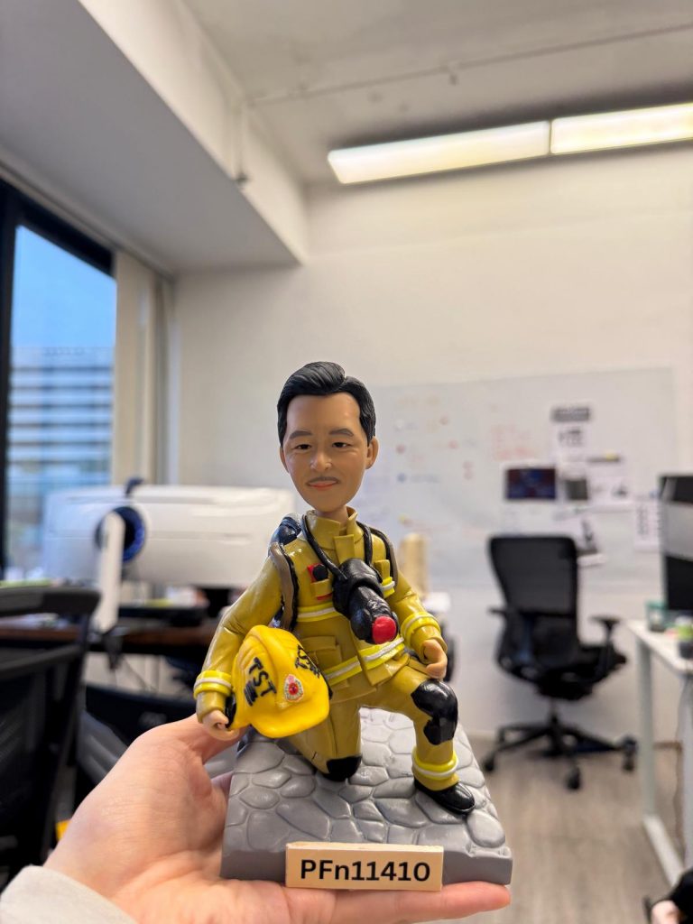 一款訂製的消防人像公仔，呈現出英雄般的單膝跪地姿勢。公仔身穿全套黃金戰衣，手持寫有「TST」的黃色頭盔，站立在石磚底座上，是一份極具紀念價值的退休禮物。