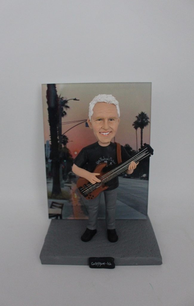 一位銀髮男士的樂手人像公仔,他身背一把低音結他,面帶微笑,站立在一塊以加州黃昏棕櫚樹街景為主題的相片背景板前。