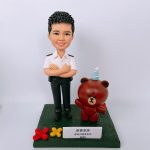 一款為香港紀律部隊人員訂製的榮休人像公仔，主角身穿白色制服，身旁站著一隻戴派對帽的Brown熊大卡通公仔作為背景配件裝飾。