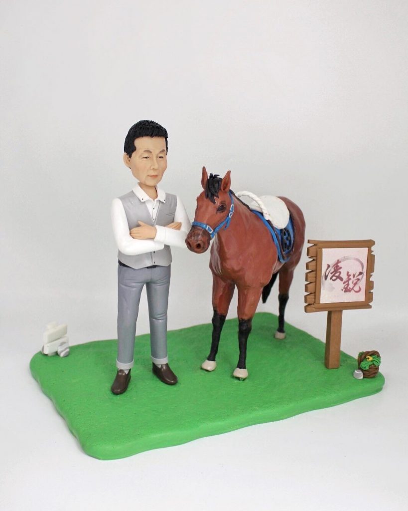 馬術主題人像公仔場景：短髮男子公仔身穿白襯衫搭灰色背心與長褲，雙手抱胸站在綠色草地底座上；身旁是一匹棕色馬，配有藍色韁繩與鞍具、白色鞍毯。右側豎立木質指示牌（附書法字樣），地面另有小型裝飾物。整體呈現馬場教學與照護氛圍，適合作為致敬馬術師的退休禮物人像公仔。