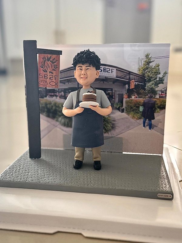 咖啡店主題人像公仔擺設：捲髮笑臉的店主穿淺灰上衣與深灰圍裙，雙手捧著巧克力分層蛋糕站在灰色質感底座上；左側有仿木製店招立牌，背板為咖啡店門面與綠植街景照片。整體呈現開業氣氛與手作風格，適合作為咖啡店開張禮物與店內展示擺設。