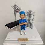 醫生人像公仔，身穿藍色手術服與口罩帽，雙手交叉站立，背景搭配手術床、手術燈及達文西手術機械臂模型。此公仔為醫生專屬客製化禮物，適合作為醫護紀念品、感謝醫生的特別禮物。