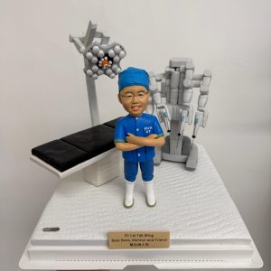 醫生人像公仔，身穿藍色手術服與口罩帽，雙手交叉站立，背景搭配手術床、手術燈及達文西手術機械臂模型。此公仔為醫生專屬客製化禮物，適合作為醫護紀念品、感謝醫生的特別禮物。