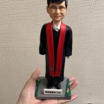 單人牧師人像公仔：黑色長袍外搭紅色聖帶，白色立領襯衫領口微露，配戴眼鏡、短黑髮，表情溫和端莊；立於灰色石板感底座，四角綠邊點綴小花，前方銘牌可客製年份與祝福文字。此人像公仔適合作為榮休、授職、周年與致謝的禮物。