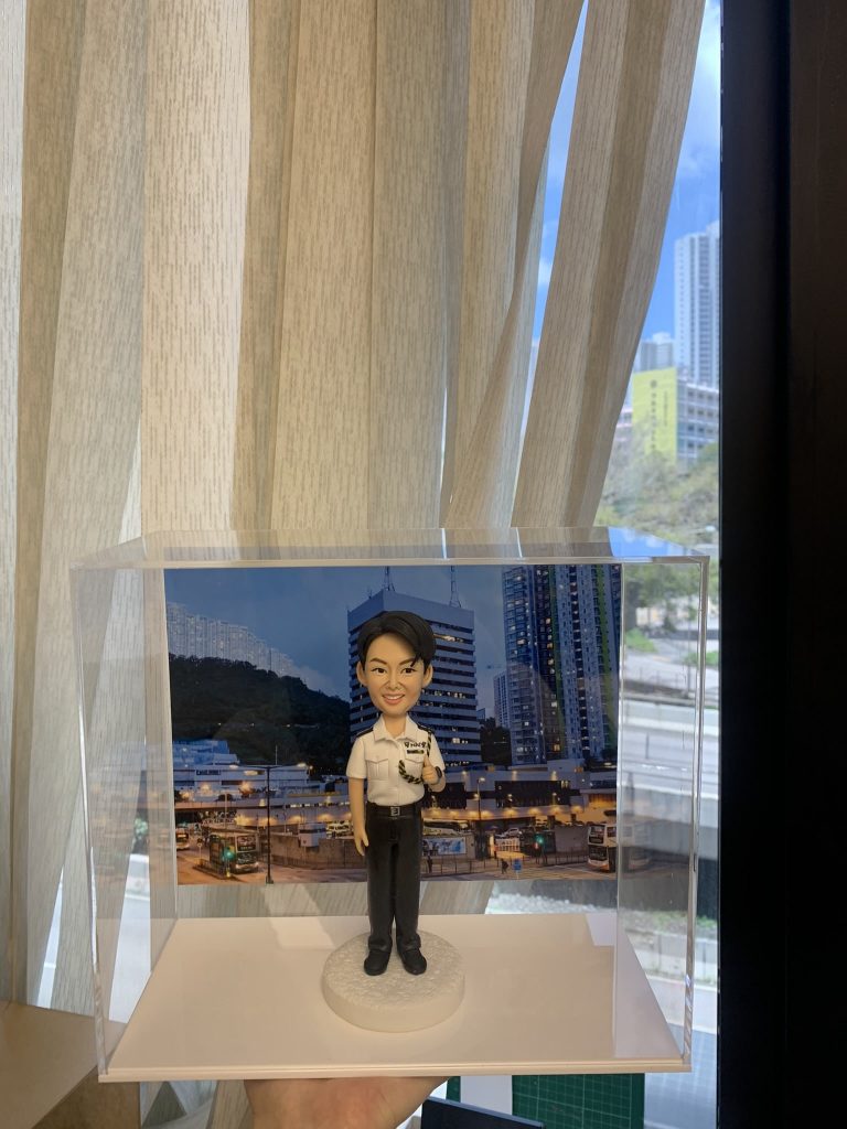 都會值勤意象融入典藏盒：本款警察人像公仔以白恤衫制服搭配通訊裝備為主視覺，臉部表情雕刻、髮絲刻線、肩章與口袋摺痕、對講機麥克風與線路、腰帶扣具等皆以手作成形並分色上色。圓形紋理底座配合城市夜景背板，凸顯專業而親和的巡邏風格。作為升遷誌慶、輪調餞別或部門致謝的送禮之選，兼具紀念與展示價值。