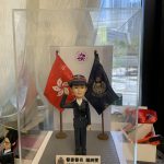 透明展示盒內的警察人像公仔：身著深藍制服與制帽，右手舉至眉間敬禮，左手自然下垂；胸前有肩帶與口袋釦飾，黑色皮鞋。人物站在灰色方形地台中央，左右各有一面旗幟：左側為紅底白花旗，右側為深藍色警徽旗。前方白底名牌印有中英對照的職級稱呼與姓名；名牌左側帶有警徽圖案。背板中央見到圓形紫色標誌，展示盒置於工作桌上，周圍有手工具與模型配件；窗外為高樓景觀。整體呈現正式、榮譽與頒贈性質，適合作為升遷、榮休或部門致意的送禮。