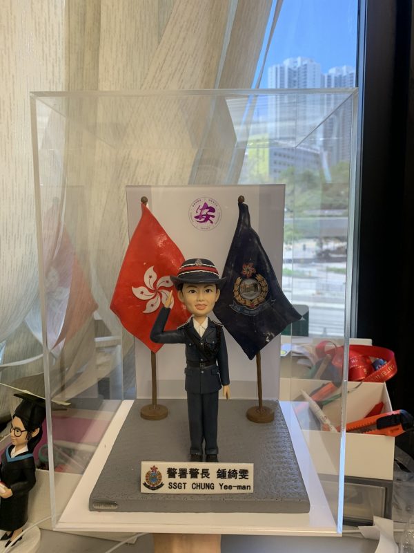 透明展示盒內的警察人像公仔：身著深藍制服與制帽，右手舉至眉間敬禮，左手自然下垂；胸前有肩帶與口袋釦飾，黑色皮鞋。人物站在灰色方形地台中央，左右各有一面旗幟：左側為紅底白花旗，右側為深藍色警徽旗。前方白底名牌印有中英對照的職級稱呼與姓名；名牌左側帶有警徽圖案。背板中央見到圓形紫色標誌，展示盒置於工作桌上，周圍有手工具與模型配件；窗外為高樓景觀。整體呈現正式、榮譽與頒贈性質，適合作為升遷、榮休或部門致意的送禮。