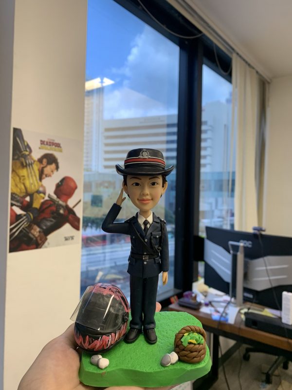 以立正敬禮定格專業形象：這款警察人像公仔以深藍制服與帽徽為主角，肩帶、口袋與鈕扣層次分明；臉部眉眼與笑意弧度、髮際線皆以手工刻線呈現。底座採草地質感，搭配塗裝安全頭盔與編織籃等客製小物，強化角色故事性。無論升遷誌慶、部門感謝或退休紀念，皆是充滿心意的送禮之選。