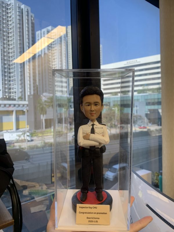 透明直立展示盒內放置一尊以「督察」職級為主題的警察人像公仔。公仔黑色短髮，神情沉穩；穿白色長袖制服襯衫，肩章與口袋翻蓋清晰，左胸可見「POLICE」字樣與名牌位置；配黑色領帶。腰間繫黑色多功能裝備腰帶，掛有對講機、套袋與工具袋；下身為深色長褲與黑色皮鞋。公仔雙臂於胸前交叉，站在黑紅相間的圓形台座上。台座前方有米金色名牌，印有英文姓名、祝賀升遷字樣與日期。展示盒由下方手掌托住，拍攝地點靠窗；窗外可見多棟高層住宅與辦公建築、道路與行道樹，藍天晴朗，玻璃上映出室內長條燈的反光，形成城市窗景與警務主題的對比。