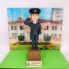 精緻手工製作的警察人像公仔（人像公仔、警察人像公仔），著深藍色正式禮服，配常服帽與帽徽、白恤衫與黑色領帶，右手舉手敬禮；胸前有斜肩皮帶與紅色禮繩，口袋、鈕扣、腰帶扣位等細節刻畫精美。公仔站在棕色台座上，前方名牌帶有警隊徽章並標示「SSGT」；背景背板為香港禮賓府外觀與花園草地，營造莊嚴典雅的典禮感。