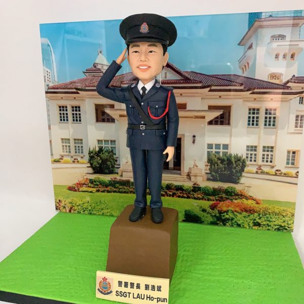 精緻手工製作的警察人像公仔（人像公仔、警察人像公仔），著深藍色正式禮服，配常服帽與帽徽、白恤衫與黑色領帶，右手舉手敬禮；胸前有斜肩皮帶與紅色禮繩，口袋、鈕扣、腰帶扣位等細節刻畫精美。公仔站在棕色台座上，前方名牌帶有警隊徽章並標示「SSGT」；背景背板為香港禮賓府外觀與花園草地，營造莊嚴典雅的典禮感。