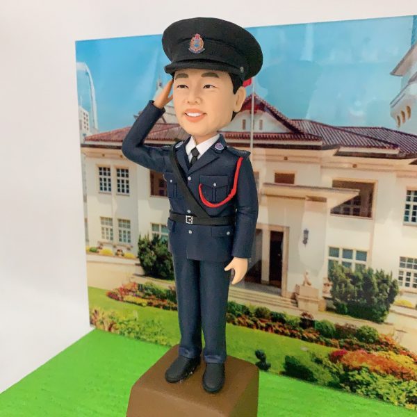 精緻手工製作的警察人像公仔（人像公仔、警察人像公仔），著深藍色正式禮服，配常服帽與帽徽、白恤衫與黑色領帶，右手舉手敬禮；胸前有斜肩皮帶與紅色禮繩，口袋、鈕扣、腰帶扣位等細節刻畫精美。公仔站在棕色台座上，前方名牌帶有警隊徽章並標示「SSGT」；背景背板為香港禮賓府外觀與花園草地，營造莊嚴典雅的典禮感。