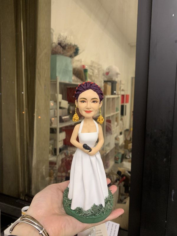 一款為擁有紫色編髮造型的女歌手訂製的人像公仔，她身穿白色長裙，配戴金色玫瑰耳環，手持咪高峰，並站立在一個綠色草地底座上。