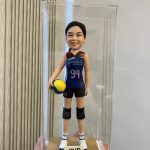 排球運動員主題人像公仔：扎馬尾的女選手身穿深藍色球衣與黑色短褲，膝部配戴護膝，右手抱著黃藍配色排球，腳穿白藍球鞋站在金色底座上；底座正面貼有「MVP」牌，整體放置於透明展示盒內，側邊可見垂直條紋牆面與手掌托起展示盒的畫面。呈現比賽頒獎後的榮耀時刻，適合作為排球比賽禮物與收藏的人像公仔。