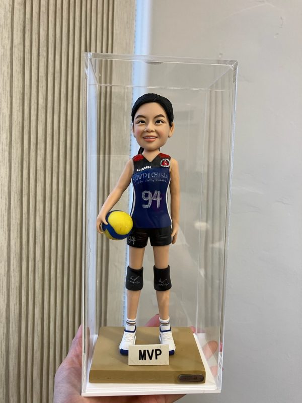 排球運動員主題人像公仔：扎馬尾的女選手身穿深藍色球衣與黑色短褲，膝部配戴護膝，右手抱著黃藍配色排球，腳穿白藍球鞋站在金色底座上；底座正面貼有「MVP」牌，整體放置於透明展示盒內，側邊可見垂直條紋牆面與手掌托起展示盒的畫面。呈現比賽頒獎後的榮耀時刻，適合作為排球比賽禮物與收藏的人像公仔。