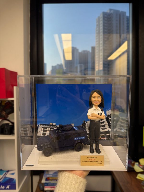 透明壓克力展示盒中，立有一尊女性「督察」主題的警察人像公仔。公仔短黑髮、微笑神情，雙臂在胸前交叉；上身穿白色短袖制服襯衫，肩章與名牌位置清晰，左胸別有對講機肩咪與連線；下身為黑色戰術長褲與黑色長靴，腰間繫有裝備腰帶。人物站於淺色紋理底座，前方設有金色名牌，印有祝賀與致謝文字。背板是一輛深藍色多用途裝甲車（POLICE標示清楚）與幾何線條感強烈的建築群；盒外環境靠近窗邊，窗外可見密集住宅高樓與藍天，室內長條燈在玻璃上留有反光。整體營造出戰術勤務與城市場景結合的紀念展示與送禮氛圍。