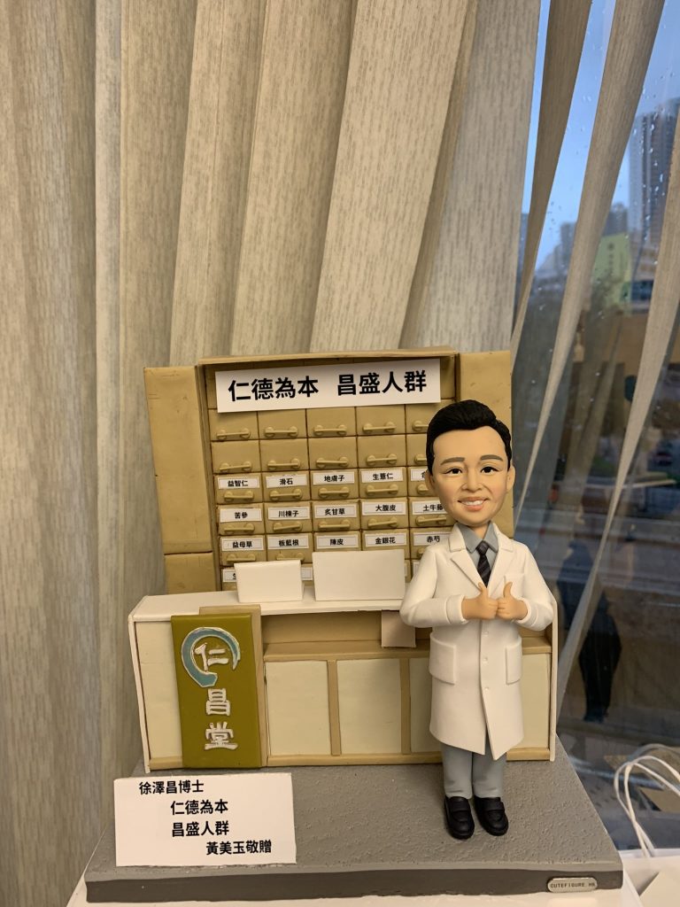 中醫師人像公仔，身穿白袍站立於中藥櫃前，背景是傳統中藥藥材抽屜櫃，上方寫有「仁德為本 昌盛人群」的牌匾。此客製化中醫公仔細節逼真，展現醫者仁心與專業精神，是送給中醫師的獨特禮物與紀念品。