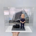 這件作品以立正合掌站姿的警察人像公仔為主角，頭部比例略大以突顯親切神情，臉型、眉眼與耳部轉折以手作雕刻呈現自然起伏，禿頂髮際、眼鏡框與唇線描繪細膩；深藍制服的口袋、肩章與縫線以細筆補色，褲管折與鞋面高光加強了質感。地台採六角石紋塑形，左側配置黑色警用裝甲車模型，含「POLICE」字樣與車頂艙口等硬派元素，與背板的政府大樓與旗幟相呼應，形成完整的警務情境。透明展示盒有效防塵並提升陳列儀式感，適合作為榮升、退休、嘉許或周年紀念的體面之選，亦非常適合同事與親友送禮；可進一步客製姓名、職級、頒贈日期與隊章背板，或增添對講機、勳表、證件夾等配件，完整展現警察人像公仔的專業與情感價值。