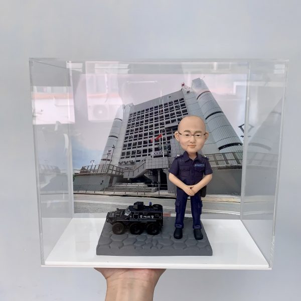 這件作品以立正合掌站姿的警察人像公仔為主角，頭部比例略大以突顯親切神情，臉型、眉眼與耳部轉折以手作雕刻呈現自然起伏，禿頂髮際、眼鏡框與唇線描繪細膩；深藍制服的口袋、肩章與縫線以細筆補色，褲管折與鞋面高光加強了質感。地台採六角石紋塑形，左側配置黑色警用裝甲車模型，含「POLICE」字樣與車頂艙口等硬派元素，與背板的政府大樓與旗幟相呼應，形成完整的警務情境。透明展示盒有效防塵並提升陳列儀式感，適合作為榮升、退休、嘉許或周年紀念的體面之選，亦非常適合同事與親友送禮；可進一步客製姓名、職級、頒贈日期與隊章背板，或增添對講機、勳表、證件夾等配件，完整展現警察人像公仔的專業與情感價值。