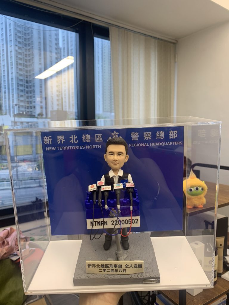 記者主題人像公仔置於透明展示盒：短髮男記者身穿深色西裝打領帶，站在藍色採訪背板前，面前排放多支新聞麥克風與採訪牌，腳下灰色台座印有姓名與日期標牌。背景可見室內窗景與高樓。整體呈現專業採訪場景，適合作為記者人像公仔榮休禮物。