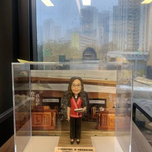 單人牧師人像公仔置於透明展示盒中：角色戴眼鏡、微笑神情，穿黑色服飾搭配紅色聖帶與白色牧領，手持書本；背景為教堂講台與木質裝飾的場景板，底座前方附有金色銘牌可刻經文或祝福語。整體精緻，適合作為教會授職、周年、榮休與致謝的禮物。