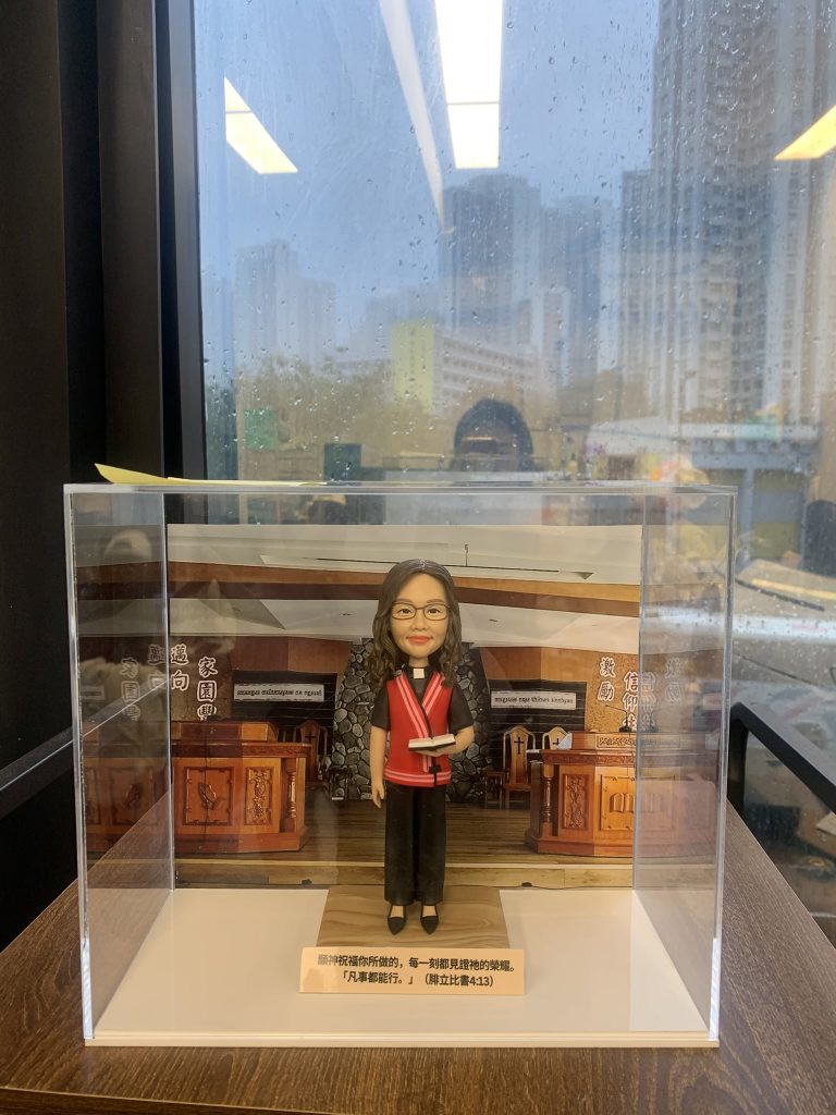 單人牧師人像公仔置於透明展示盒中：角色戴眼鏡、微笑神情，穿黑色服飾搭配紅色聖帶與白色牧領，手持書本；背景為教堂講台與木質裝飾的場景板，底座前方附有金色銘牌可刻經文或祝福語。整體精緻，適合作為教會授職、周年、榮休與致謝的禮物。