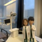 兩位身穿白袍的醫生人像公仔，站立於醫療場景模型前，背景包含電腦顯示屏與手術燈。公仔細節精緻，展現專業醫護形象，腳踩藍色鞋套，象徵無菌醫療環境。底座刻有「于春紅醫生」字樣，這款專屬定製醫護公仔極具收藏與送禮價值。