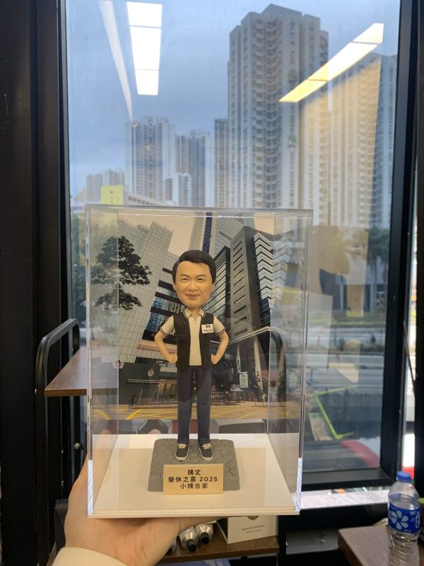 透明直立展示盒中擺放一尊便衣風格的「偵緝警員」主題人像公仔。人物黑色短髮、微笑表情，雙手插腰站姿輕鬆自信；上身穿白色短袖POLO衫，外搭黑色無袖戰術背心，左胸貼有「POLICE」與名牌識別貼；下身為深色長褲與黑色便鞋。腳下是灰色六角石鋪面造型台座，前方金色名牌以繁體中文寫有姓名與「榮休之樂 2025」等致意文字。背板為十字路口前的高樓商業區外牆與玻璃帷幕街景，地面見黃格線路口與行人道；展示盒由下方手掌托起，外部環境靠窗，窗外可見高密度住宅大廈與道路車流，窗玻璃映出室內長條燈反光與書架物件。