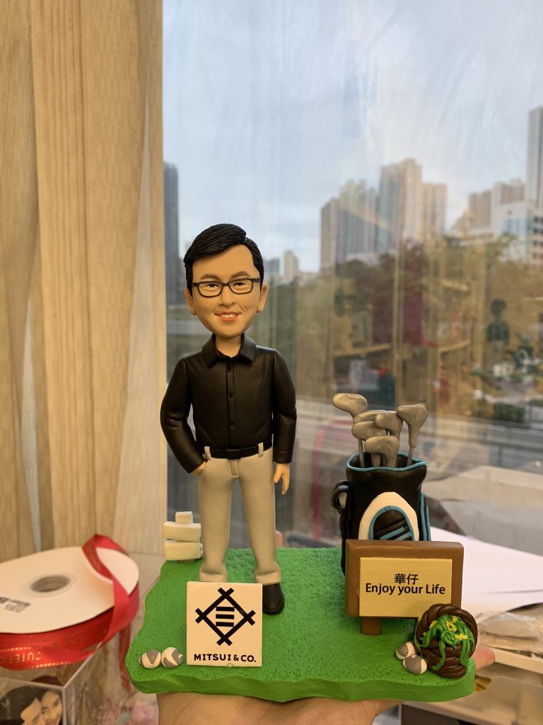 一個男同事的人像公仔站立在高爾夫主題造景模型上，身穿黑色襯衫與灰色長褲，旁邊有高爾夫球袋、球具與標語牌，牌子上寫著「華仔 Enjoy your Life」。