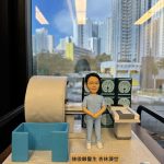 透明展示盒內的人像公仔：一位身穿淺藍色醫療制服的男醫生雙手合於腹前，身旁擺放著一台大型醫療影像檢查設備（MRI 磁力共振掃描機），後方螢幕顯示多張腦部斷層掃描影像，整體場景呈現放射科醫生的臨床專業環境。