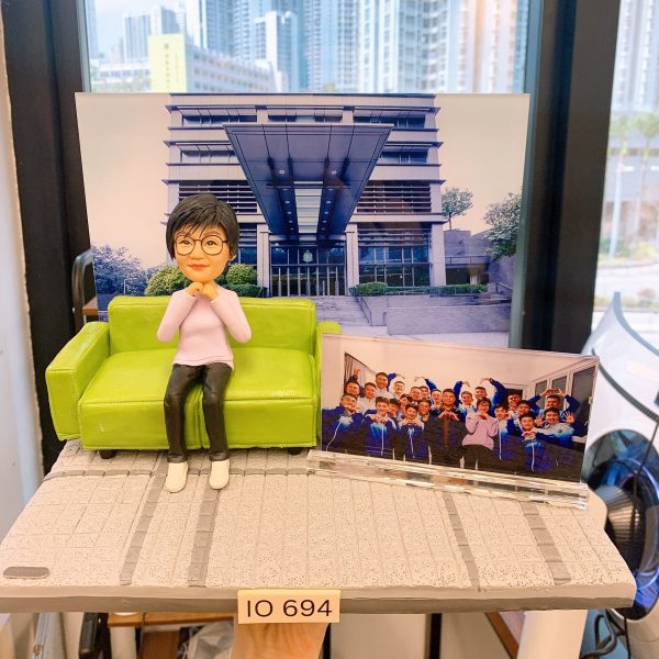 一款以警察為主題的人像公仔，身穿便服坐在綠色沙發上，身旁展示著與同袍的合照，背景是警察相關建築，底座前方印有「IO 694」字牌，象徵個人專屬身份與紀念。