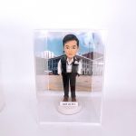 置於透明展示盒中的警察人像公仔：男性造型短髮，身著白色襯衫、深色長褲與黑色戰術背心，胸前別有「POLICE」識別章與名牌，雙手插袋站姿挺直；腳穿棕色鞋，立於白色圓形底座，前方貼有姓名縮寫標籤「DSIP LAI W.L.」。背景為戶外部門建築與廣場圖像，地面有黃色標線與旗桿，營造執勤場景氛圍。整體在純白環境中拍攝，突顯公仔細節與升職紀念意義。