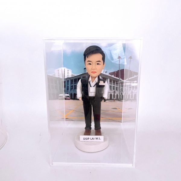 置於透明展示盒中的警察人像公仔：男性造型短髮，身著白色襯衫、深色長褲與黑色戰術背心，胸前別有「POLICE」識別章與名牌，雙手插袋站姿挺直；腳穿棕色鞋，立於白色圓形底座，前方貼有姓名縮寫標籤「DSIP LAI W.L.」。背景為戶外部門建築與廣場圖像，地面有黃色標線與旗桿，營造執勤場景氛圍。整體在純白環境中拍攝，突顯公仔細節與升職紀念意義。
